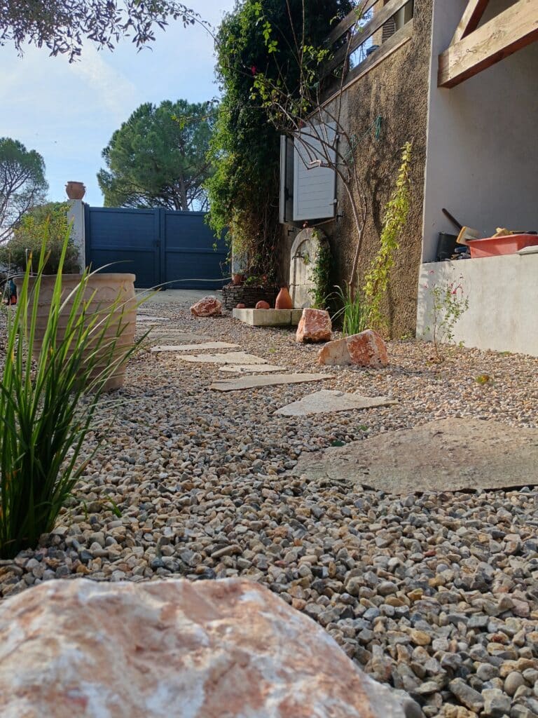 Allée de jardin en gravier avec pas japonais menant à un portail bleu, fontaine en pierre et plantes grimpantes.