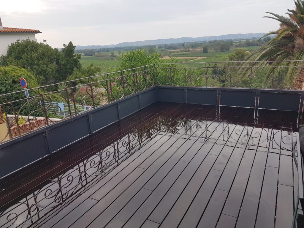 Balcon avec terrasse en bois sombre mouillée et garde-corps en fer forgé surplombant des vignes et collines verdoyantes.