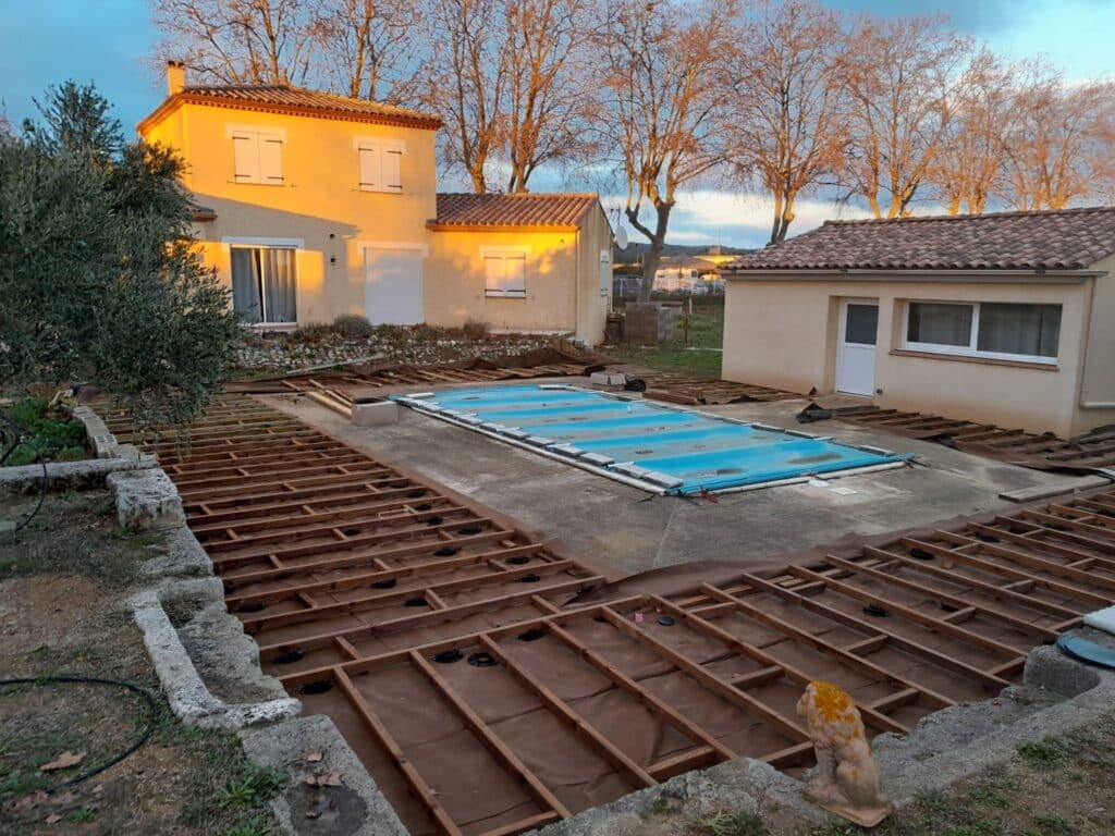 Structure de terrasse bois en construction autour d'une piscine couverte avec maison jaune.