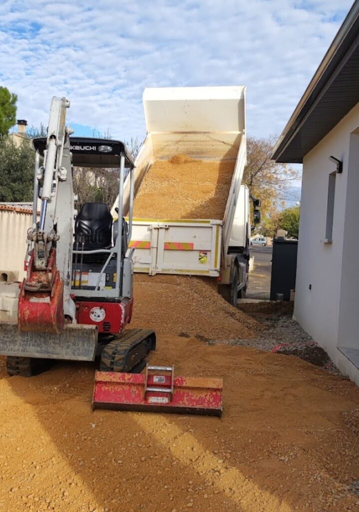 Camion déchargeant du gravier jaune près d'une mini-pelle Takeuchi sur un chantier de terrassement.